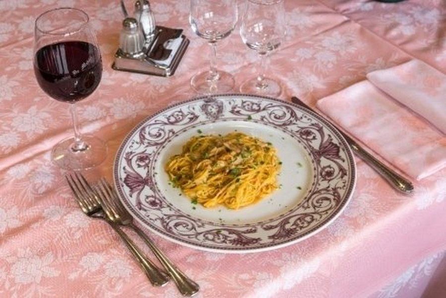 Elegante apparecchiatura della tavola con pasta su un piatto decorato, un bicchiere di vino rosso e posate su una tovaglia rosa.