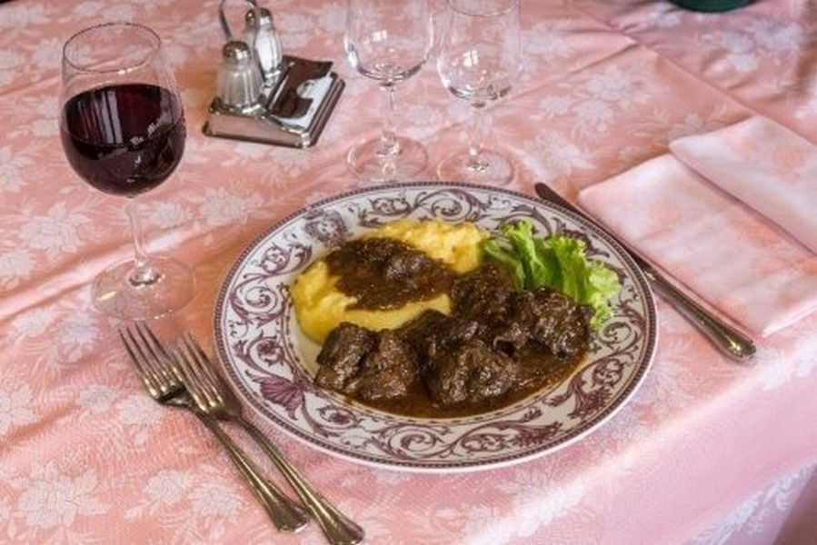 Cena a base di bistecca con purè di patate e verdure su una tovaglia rosa, accompagnata da un bicchiere di vino rosso.