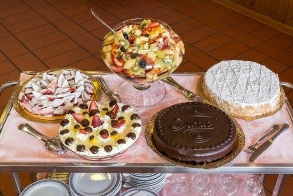 Su un tavolo si trovano diverse torte decorate, tra cui torte al cioccolato e torte con frutta e zucchero a velo.