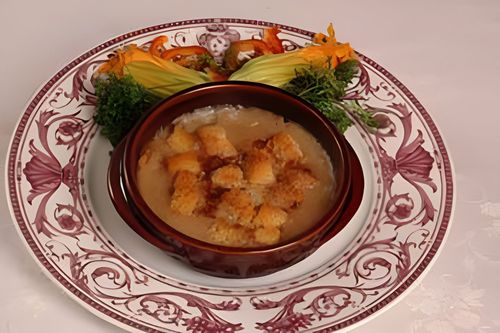 Una ciotola di zuppa cremosa con crostini su un piatto decorato con motivi floreali, guarnita con prezzemolo e fette di pomodoro.