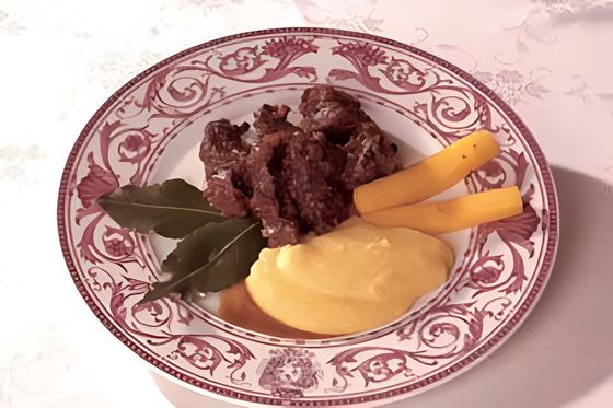 Piatto con carne grigliata, foglie di alloro, salsa gialla e bastoncini di carota su un piatto decorativo con bordo rosa.
