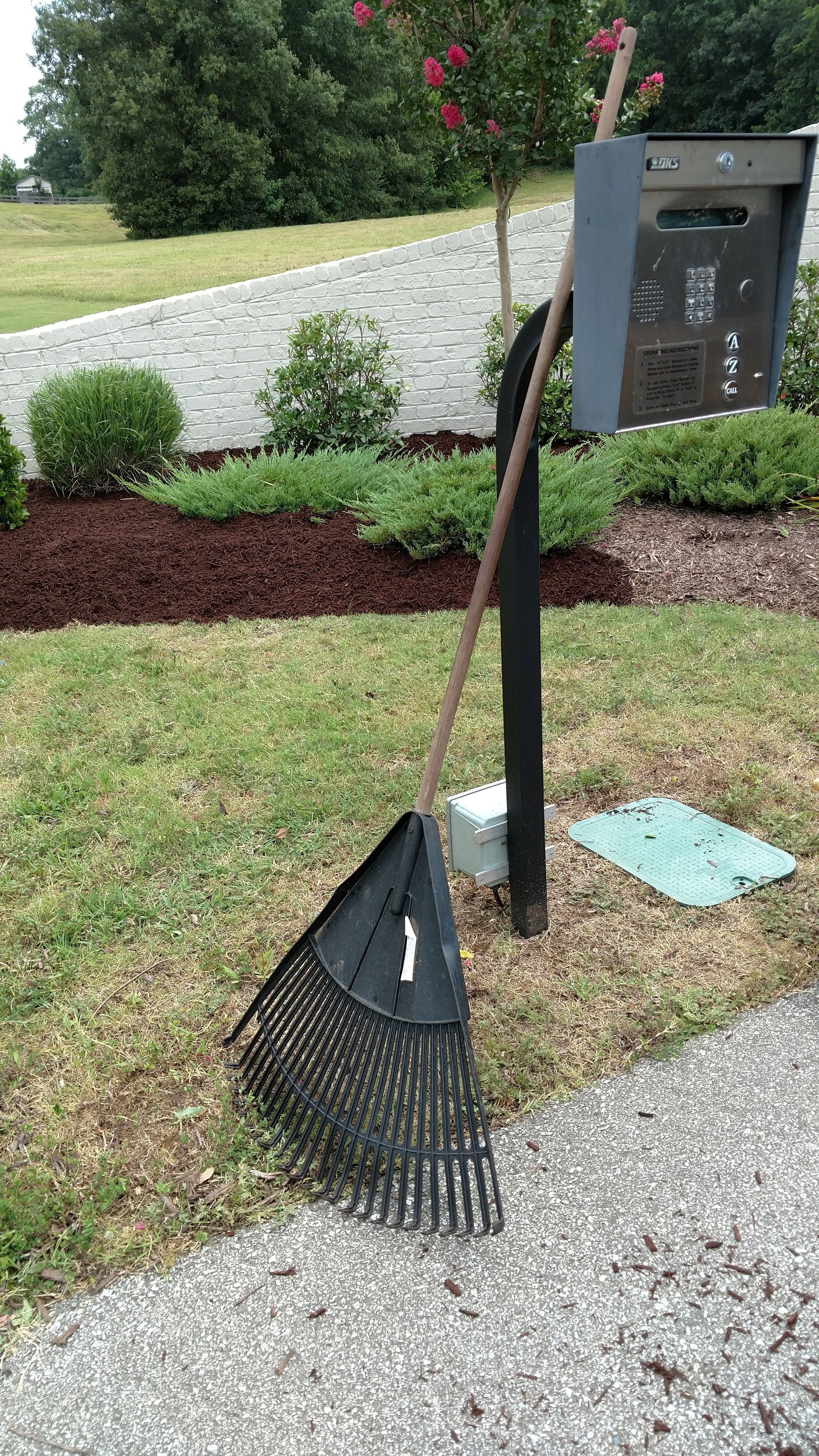 Leaf blower and trash can mounted on a post beside a sidewalk and grass in a landscaped yard.