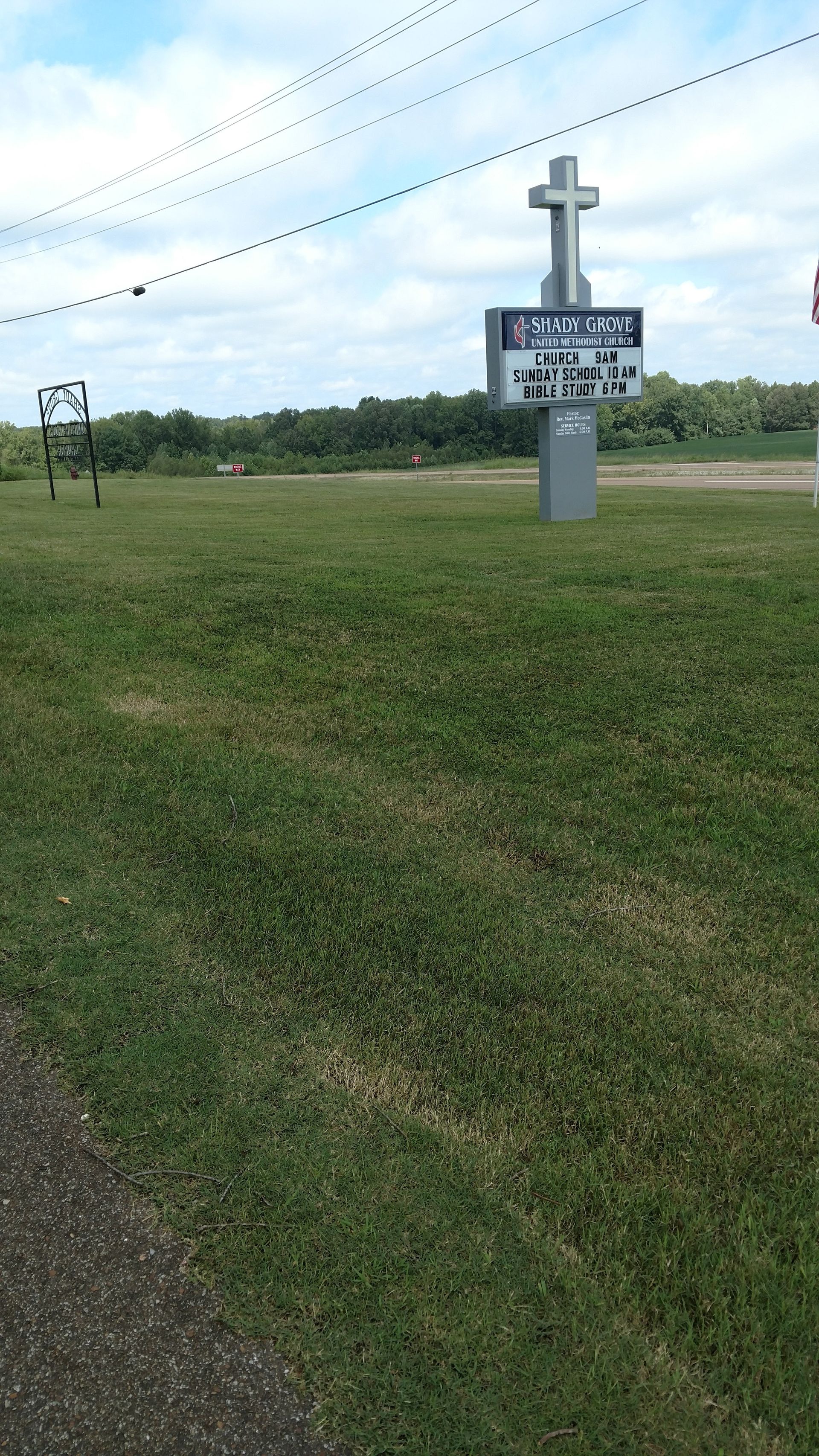 Rural roadside sign topped with a white cross beside a grassy field under power lines
