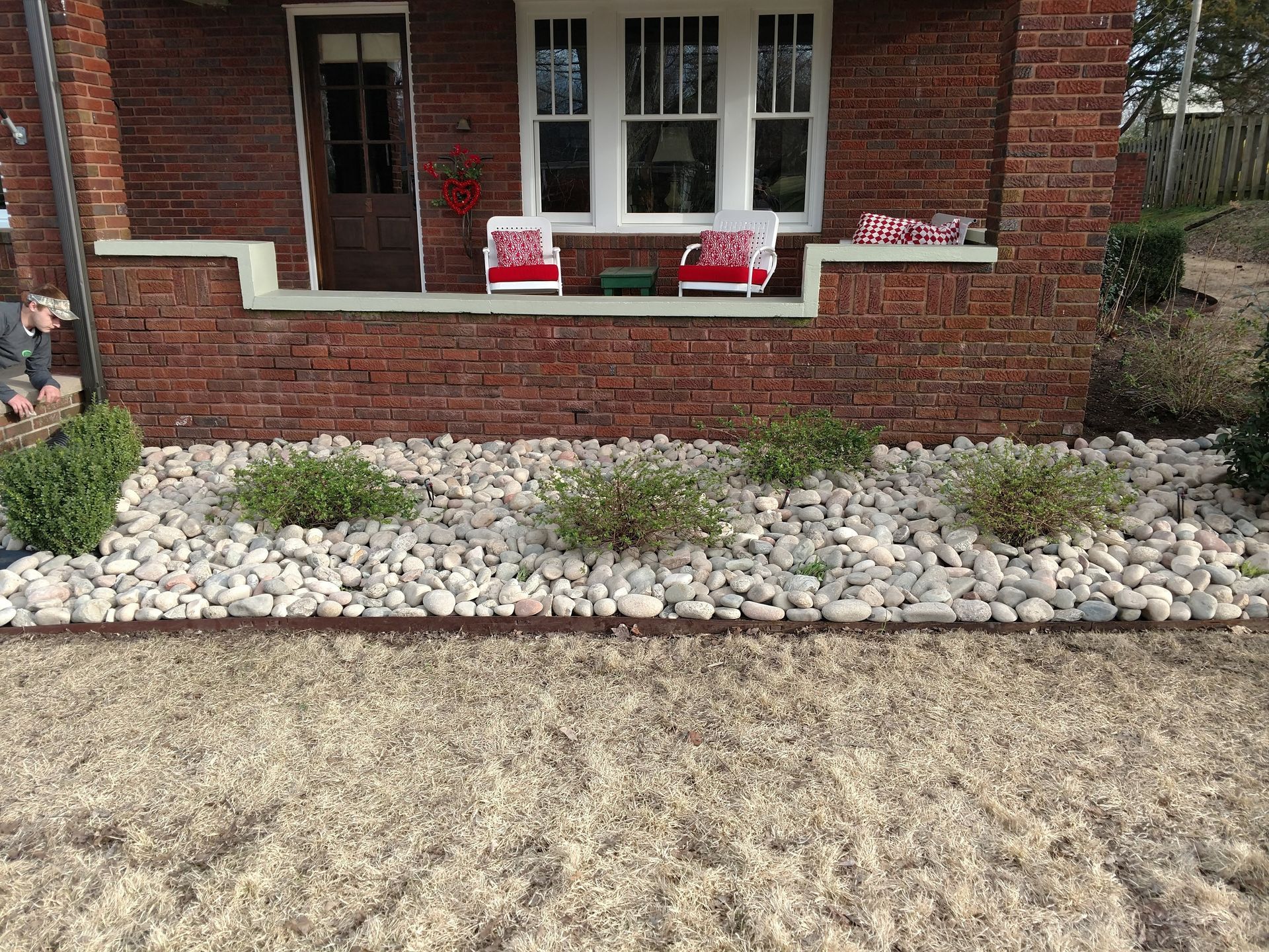 Brick house front porch with two red chairs, white stone landscaping, and a dry yellow lawn.