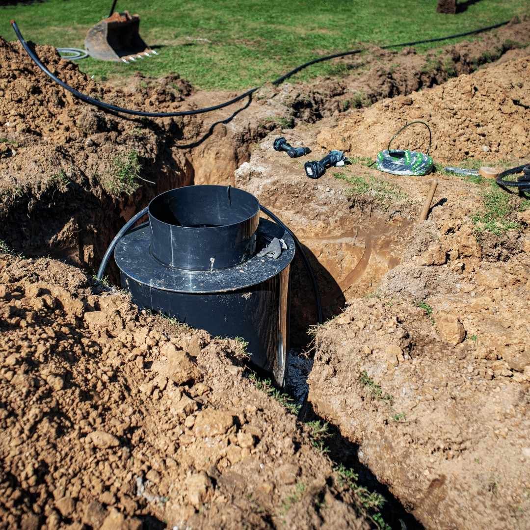 Black plastic pipe protruding from a dirt trench in a grassy yard, with tools nearby.
