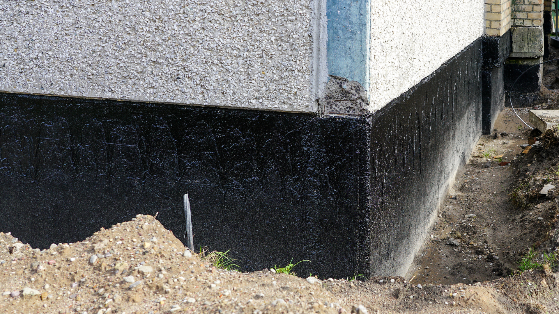 House corner with black waterproofing along the foundation and dirt trench beside it.