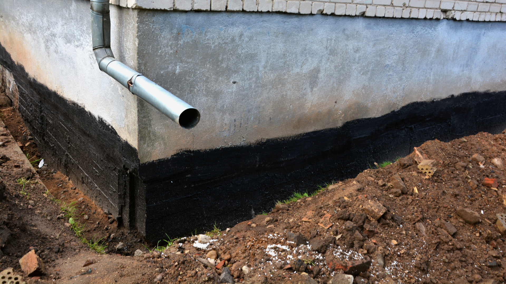 White drain pipe protruding from a concrete wall with black waterproofing at the base beside dirt ground