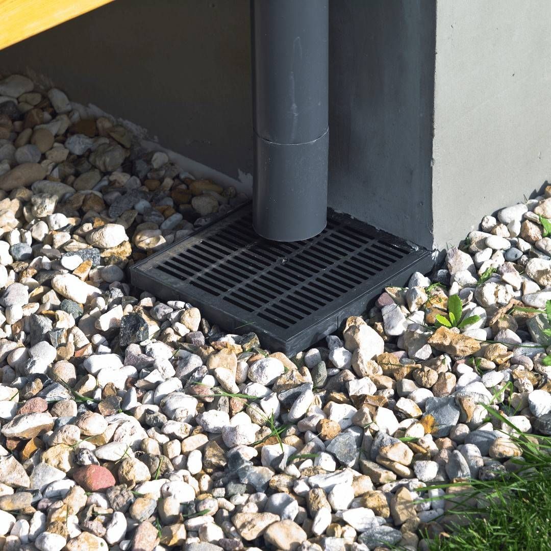 White downspout pouring water beside a brick wall onto dark mulch.