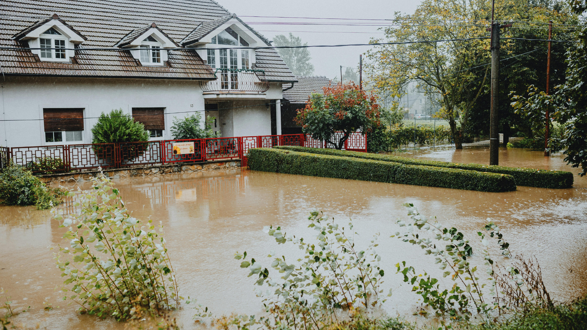 Flooded residential street with a white house, hedge, and muddy water covering the front yard