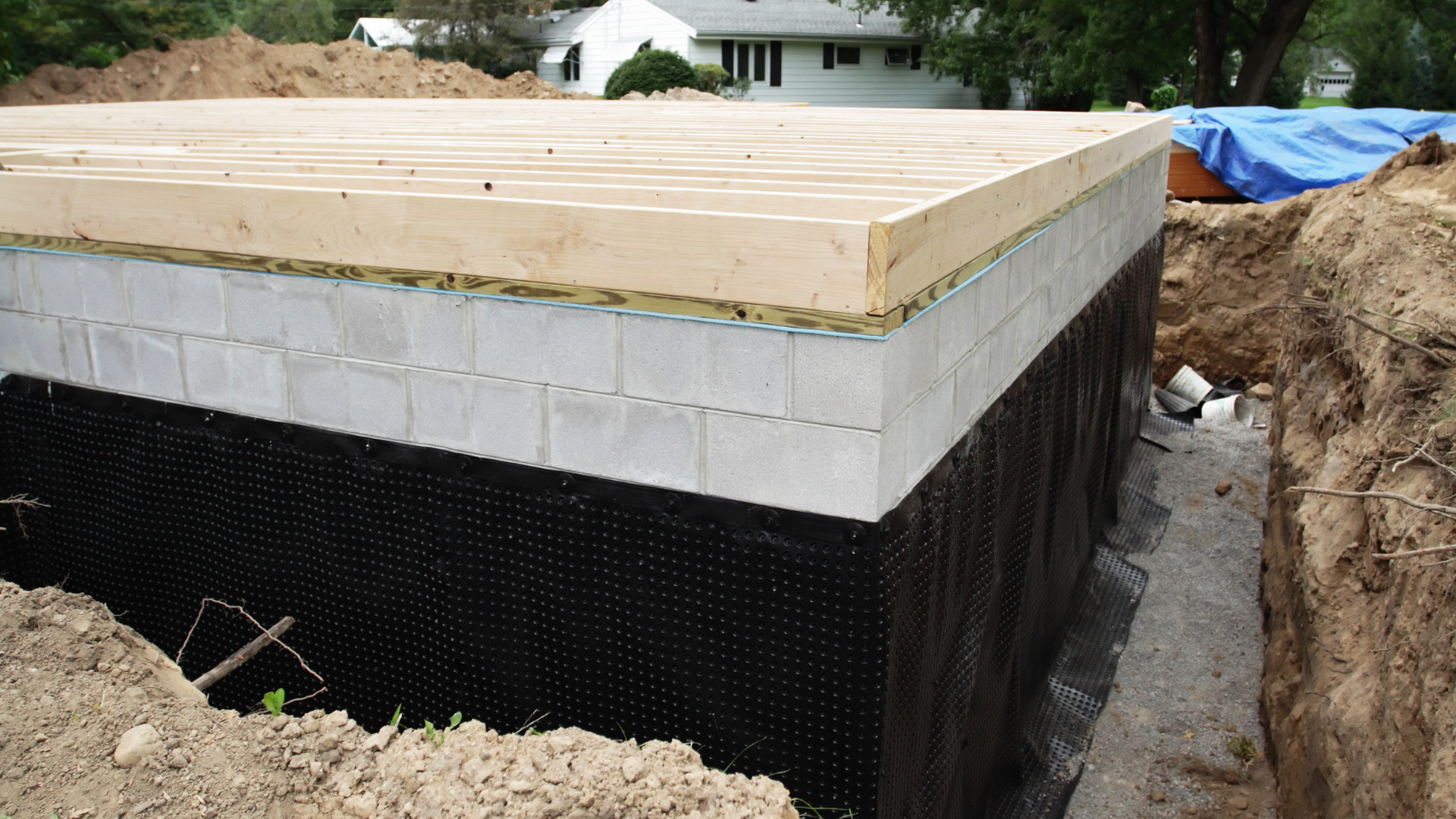 Foundation wall under construction with black waterproofing, cinder blocks, and plywood framing on top.
