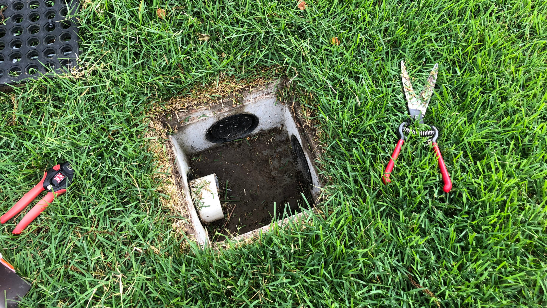 Open utility access box in grass, with red-handled tools on either side