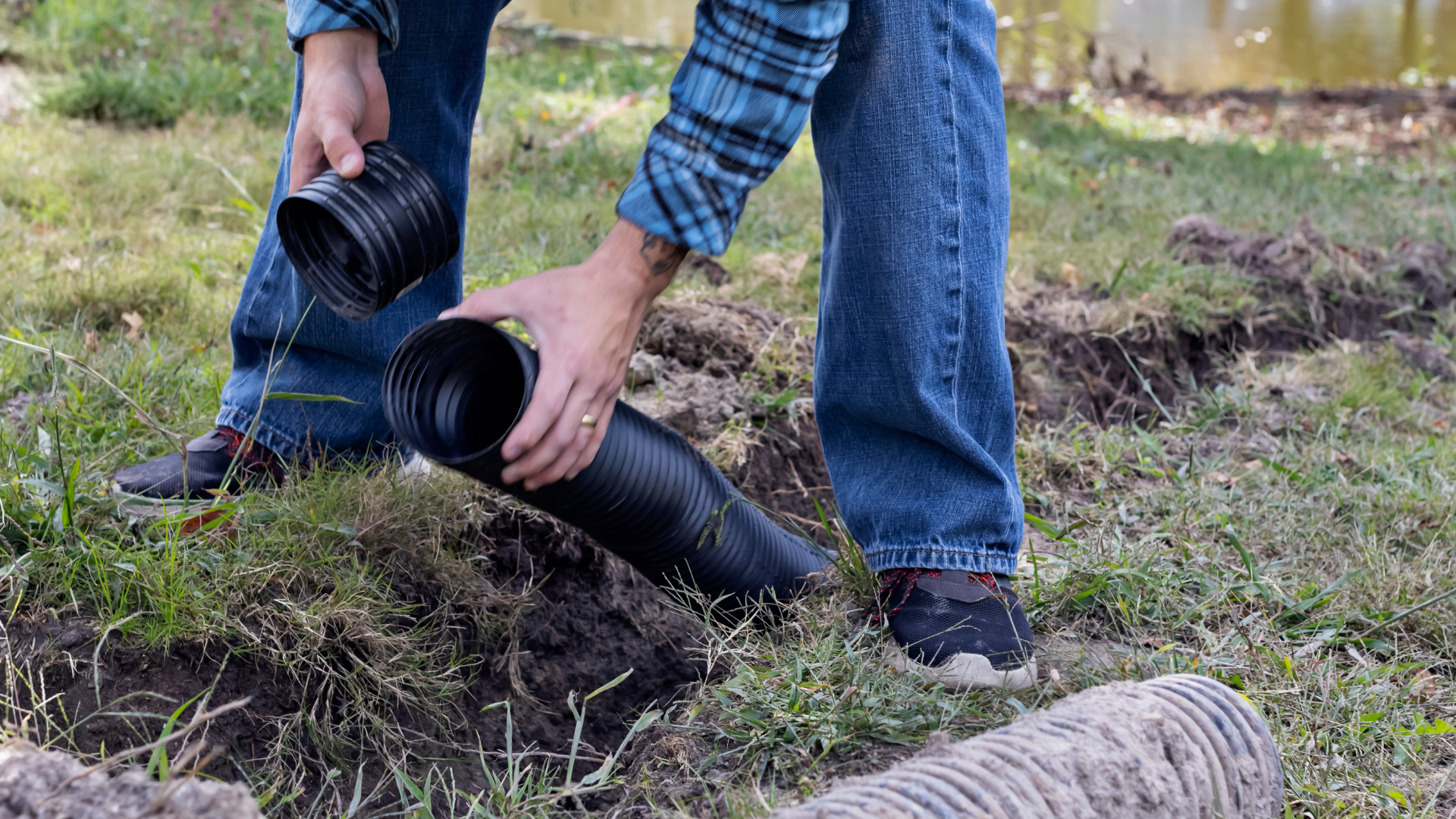 Hands in plaid sleeves placing black irrigation pipe on grassy ground near water.