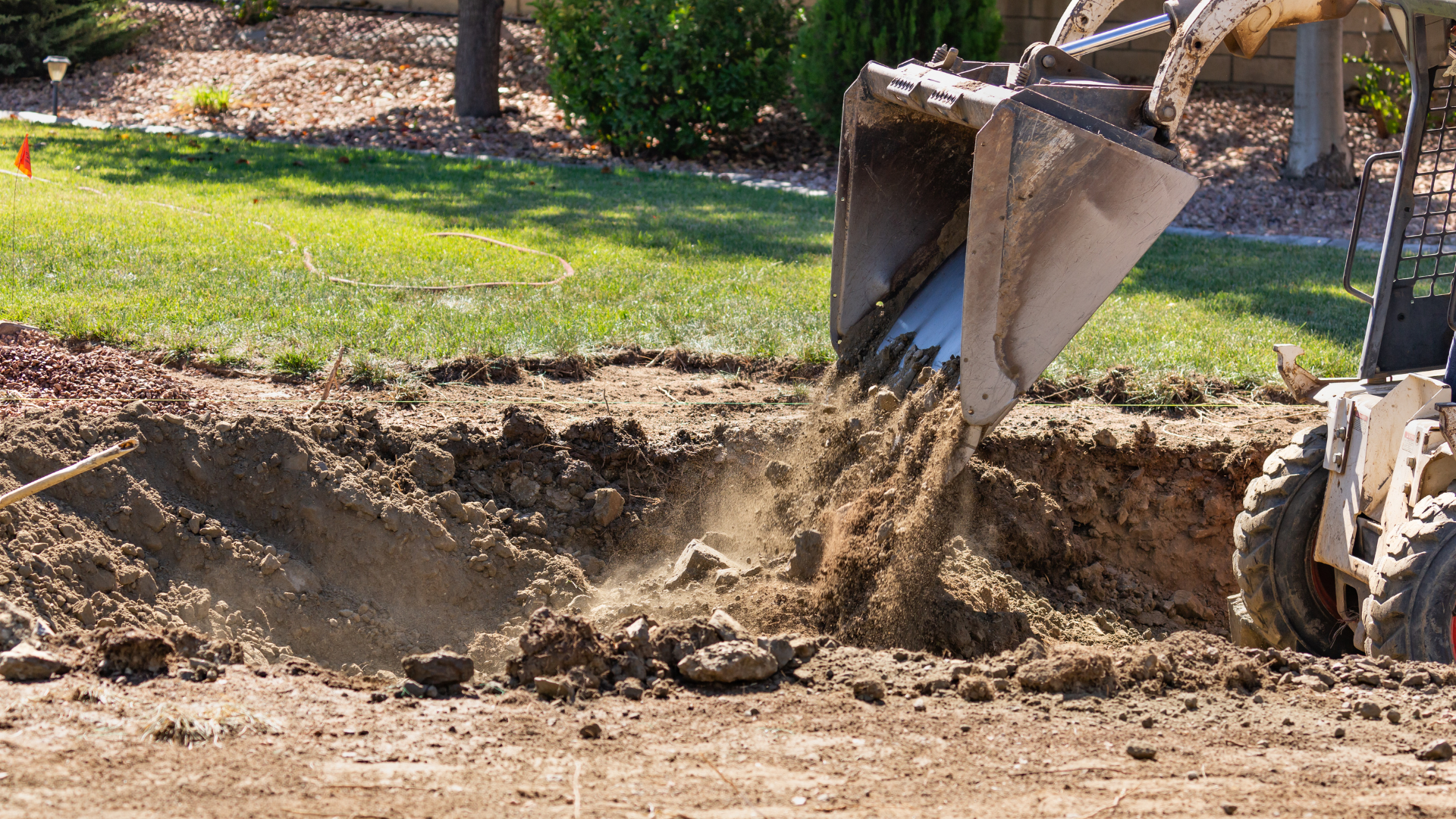 Excavator dumping dirt into a trench in a grassy yard construction site