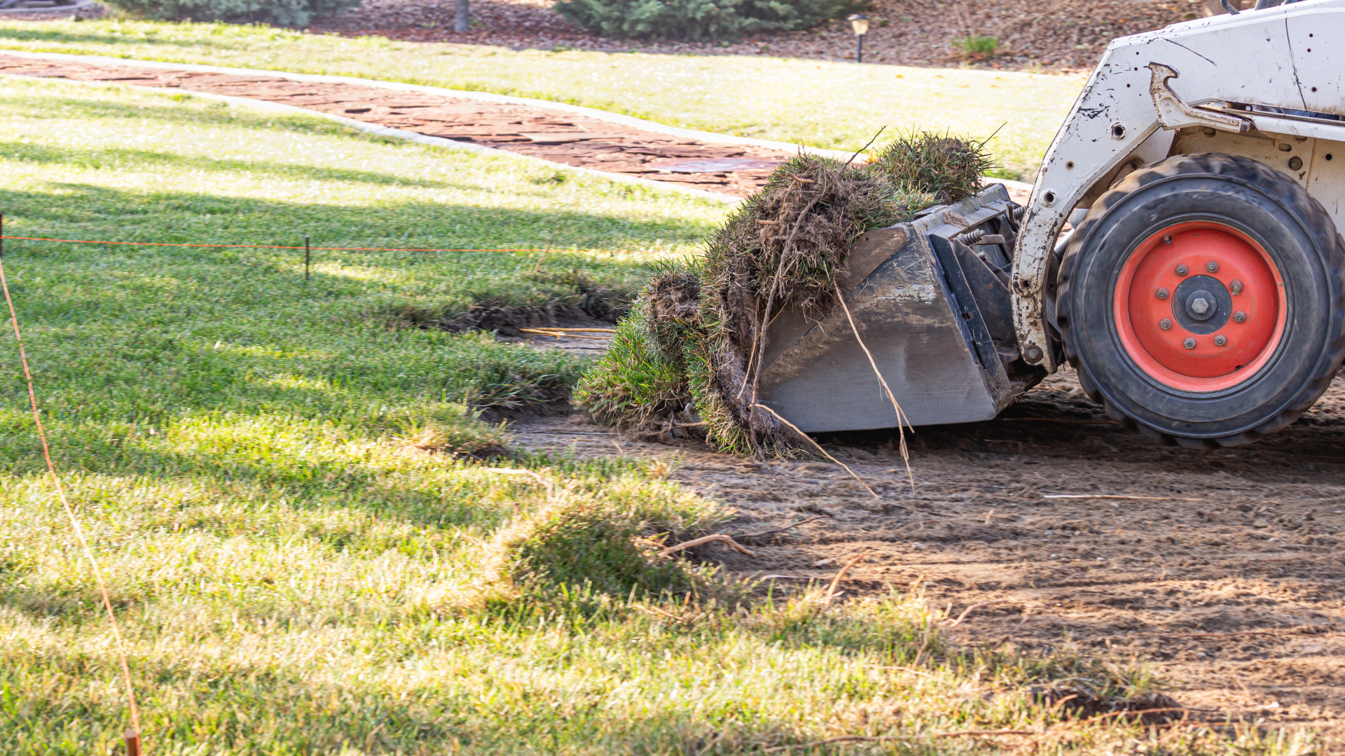 Skid steer loader clearing dirt on a grassy yard, with a pile of soil near the bucket and wheel visible