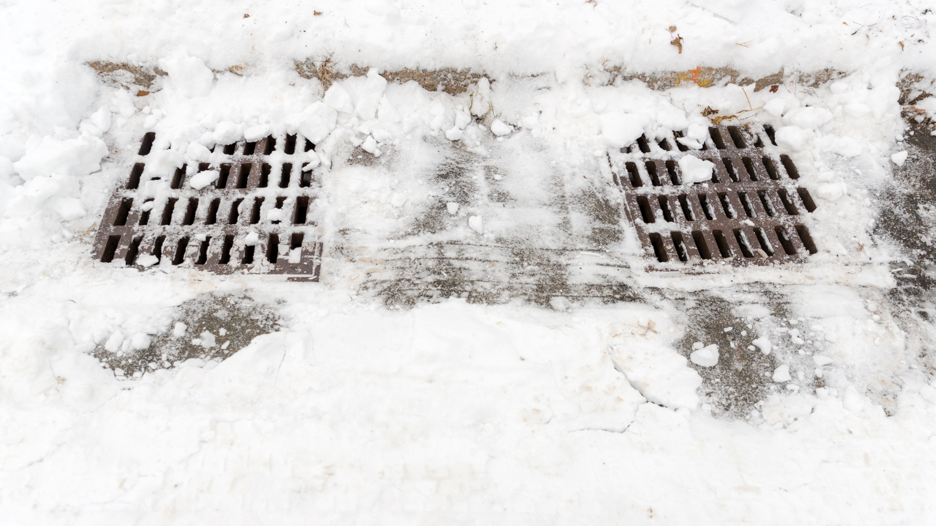 Snow-covered sidewalk drains with two metal grates visible through the snow.