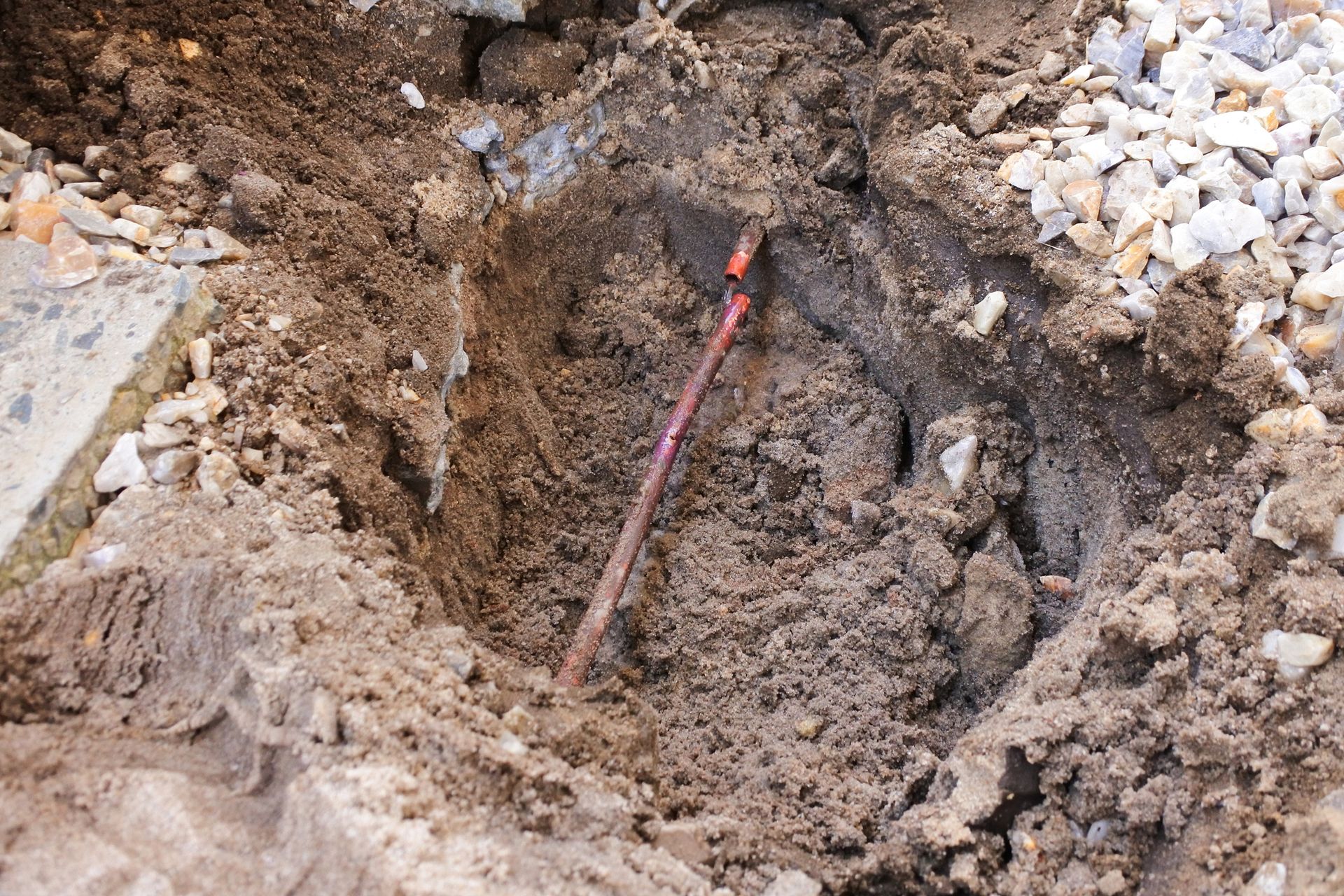 Hole dug in the dirt with a copper pipe visible. Surrounding stone, cement and loose dirt.