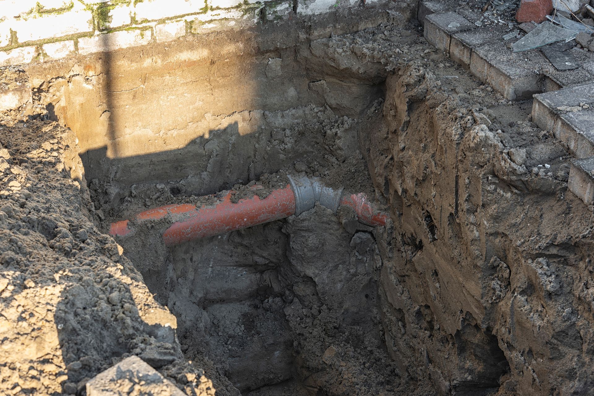A trench dug next to a brick wall exposes orange and gray drainage pipes, with surrounding dirt.