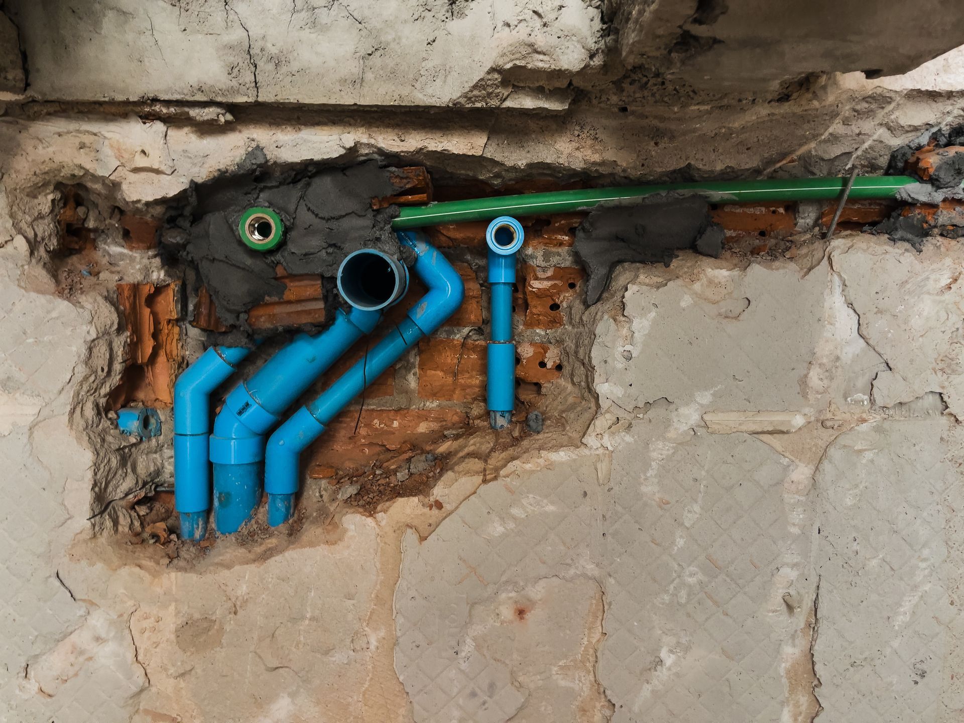 Blue PVC plumbing pipes emerging from a wall, with a green pipe and exposed brick visible.