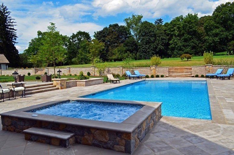 Backyard pool with a stone hot tub, blue lounge chairs, and lush green trees under a cloudy sky