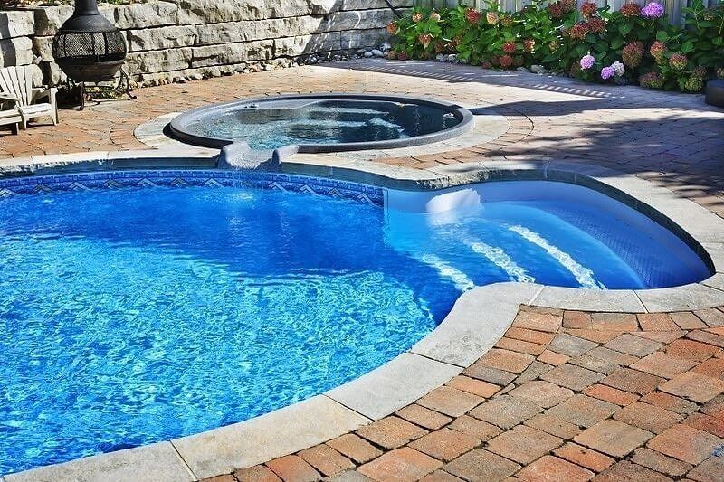 Blue backyard pool with stone hot tub, curving slide, and brick patio surround
