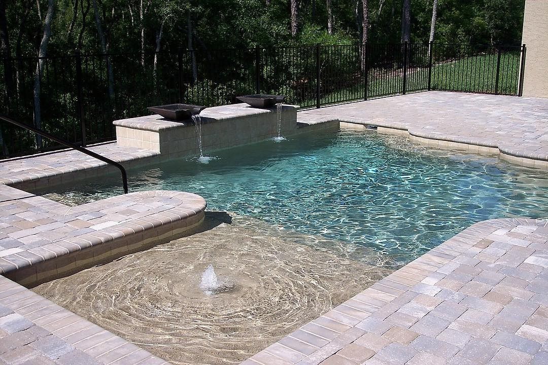 Backyard pool with a shallow pebble beach entry, clear blue water, and stone decking surrounded by trees
