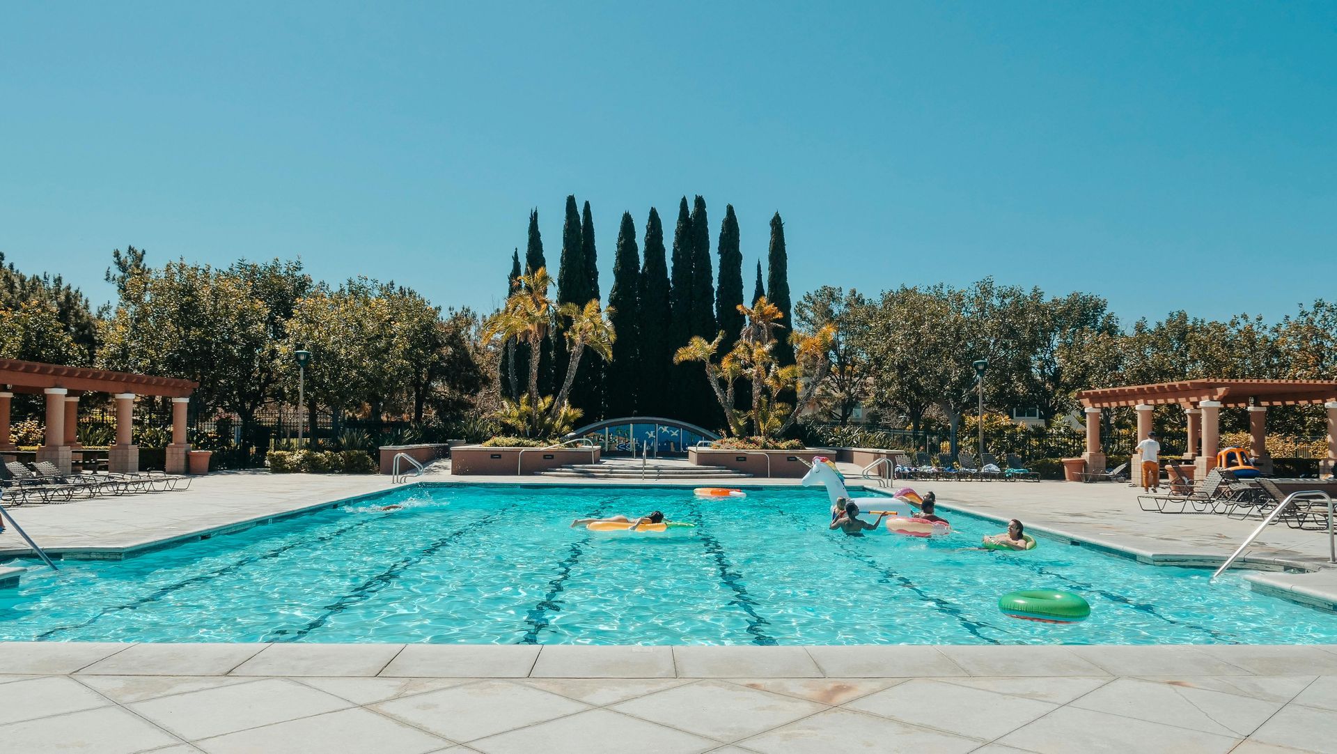 Outdoor resort pool with turquoise water, lounge chairs, and tall cypress trees under a clear blue sky