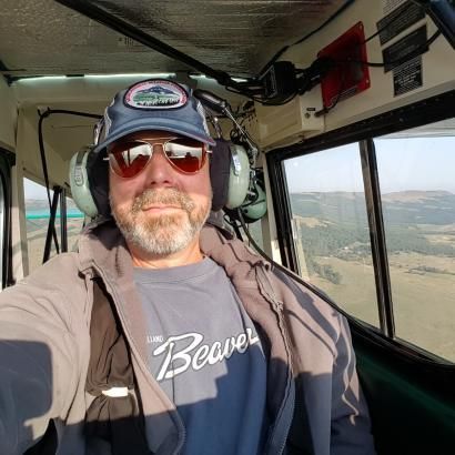 A man wearing a hat and sunglasses is taking a selfie in the cockpit of a helicopter.