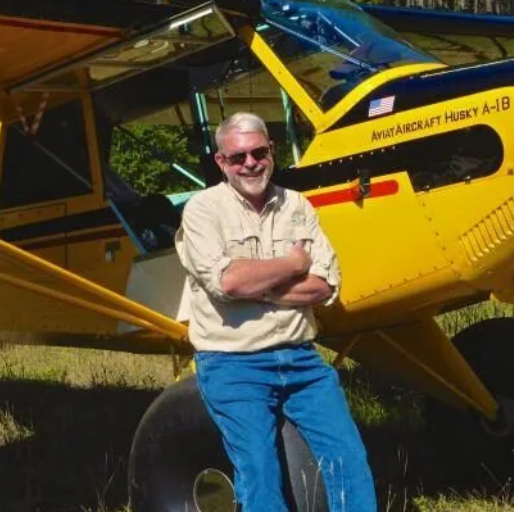 Man in hat and sunglasses in front of a small white airplane on a white plane. Blue sky.