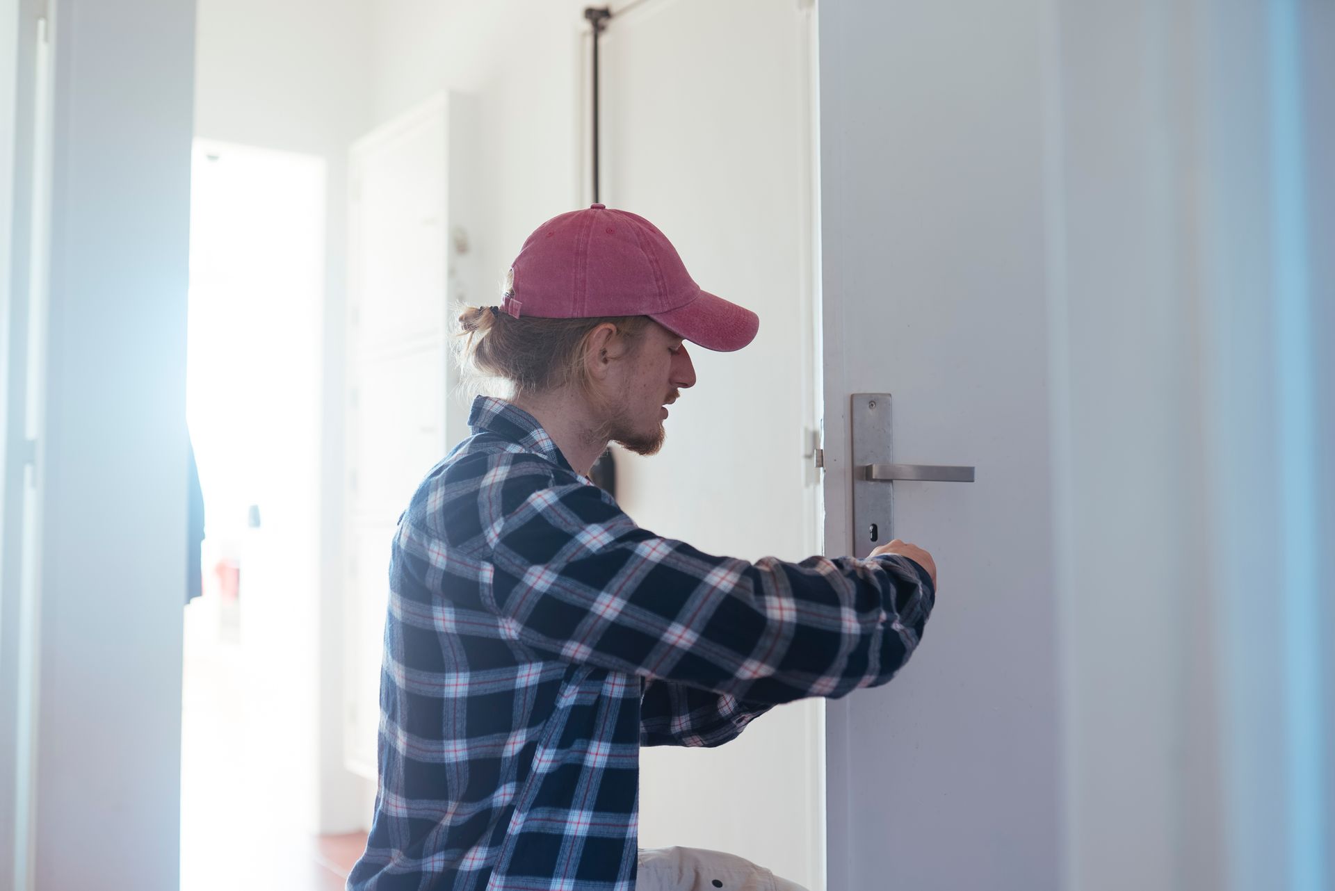 Person in plaid shirt and red cap installing a door handle in a brightly lit hallway. Person in plaid shirt and red cap installing a door handle in a brightly lit hallway.