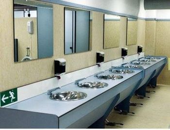 Bagno pubblico con più lavandini, specchi e dispenser di sapone. Pareti beige, banconi blu e segnaletica per l'uscita di emergenza.