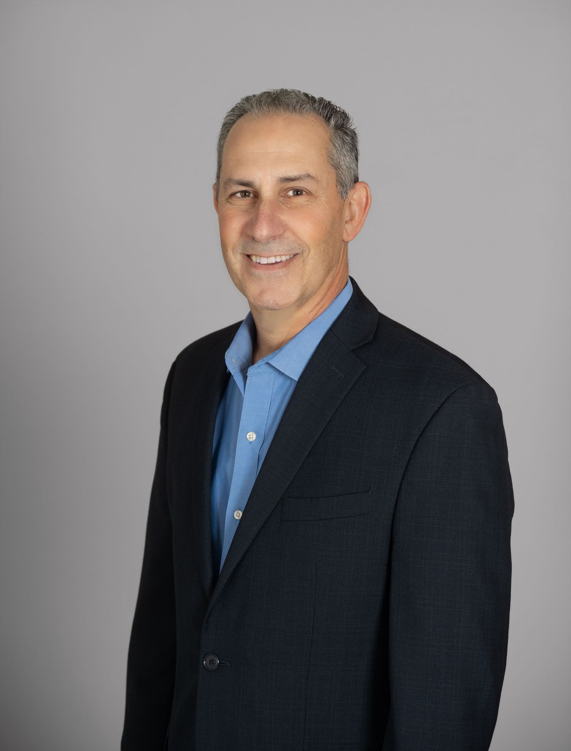 Man in blue shirt and navy blazer smiles, standing against a gray backdrop.