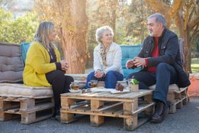 Three people sitting outdoors, talking. Wooden pallet furniture, table with drinks, sunny setting.