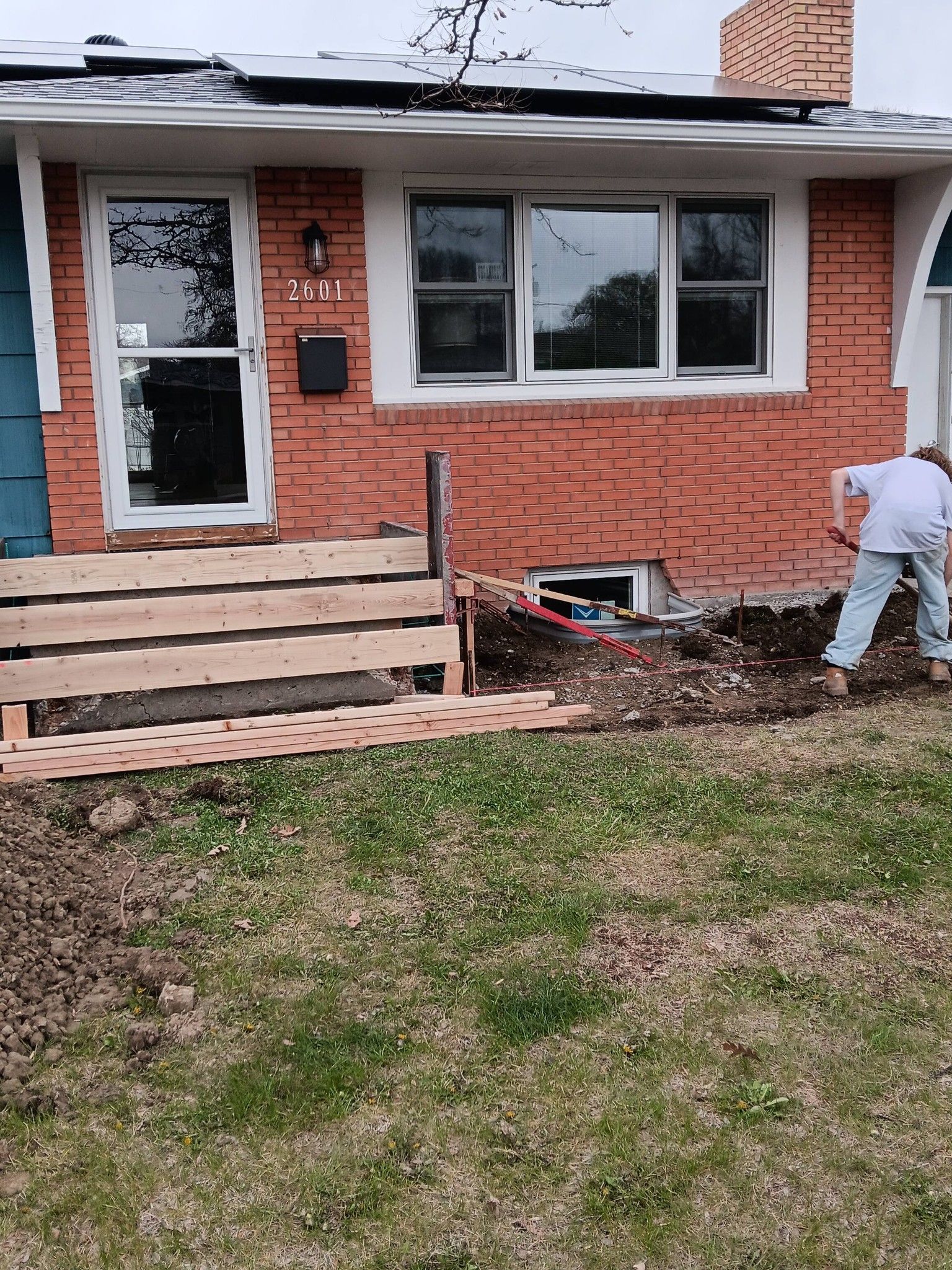 Brick house front porch under construction, with a worker beside the steps and a small white dog by the window.