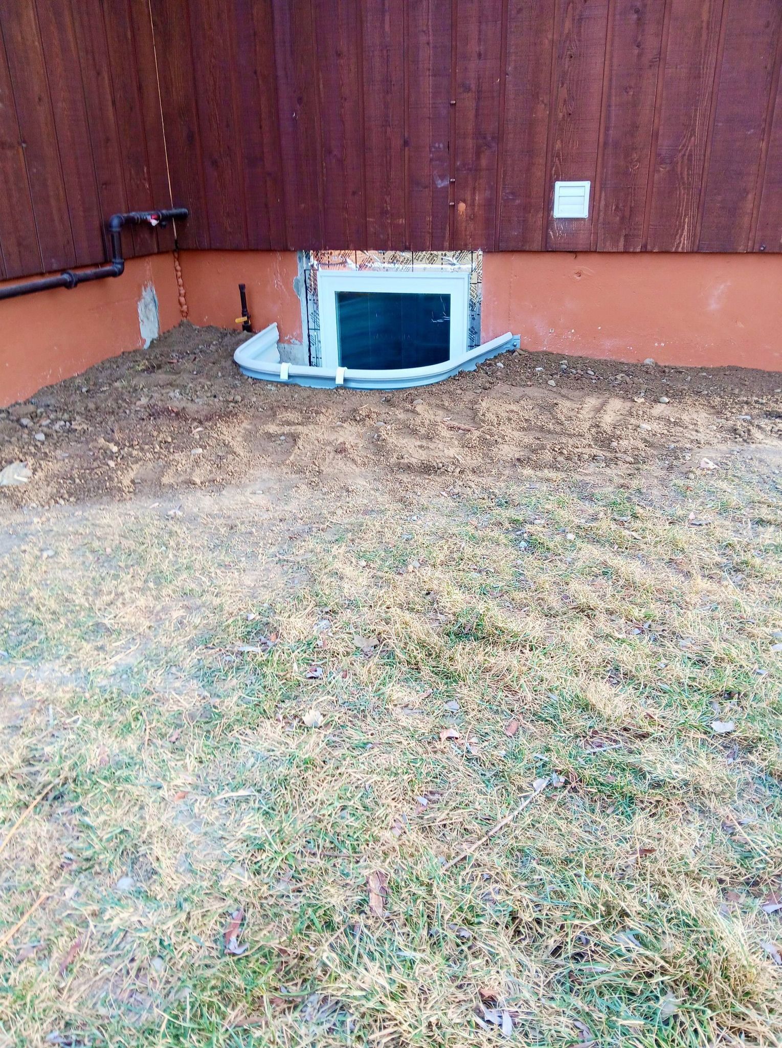 Backyard crawlspace window with white trim in a brown wooden fence, surrounded by dry grass and leaves