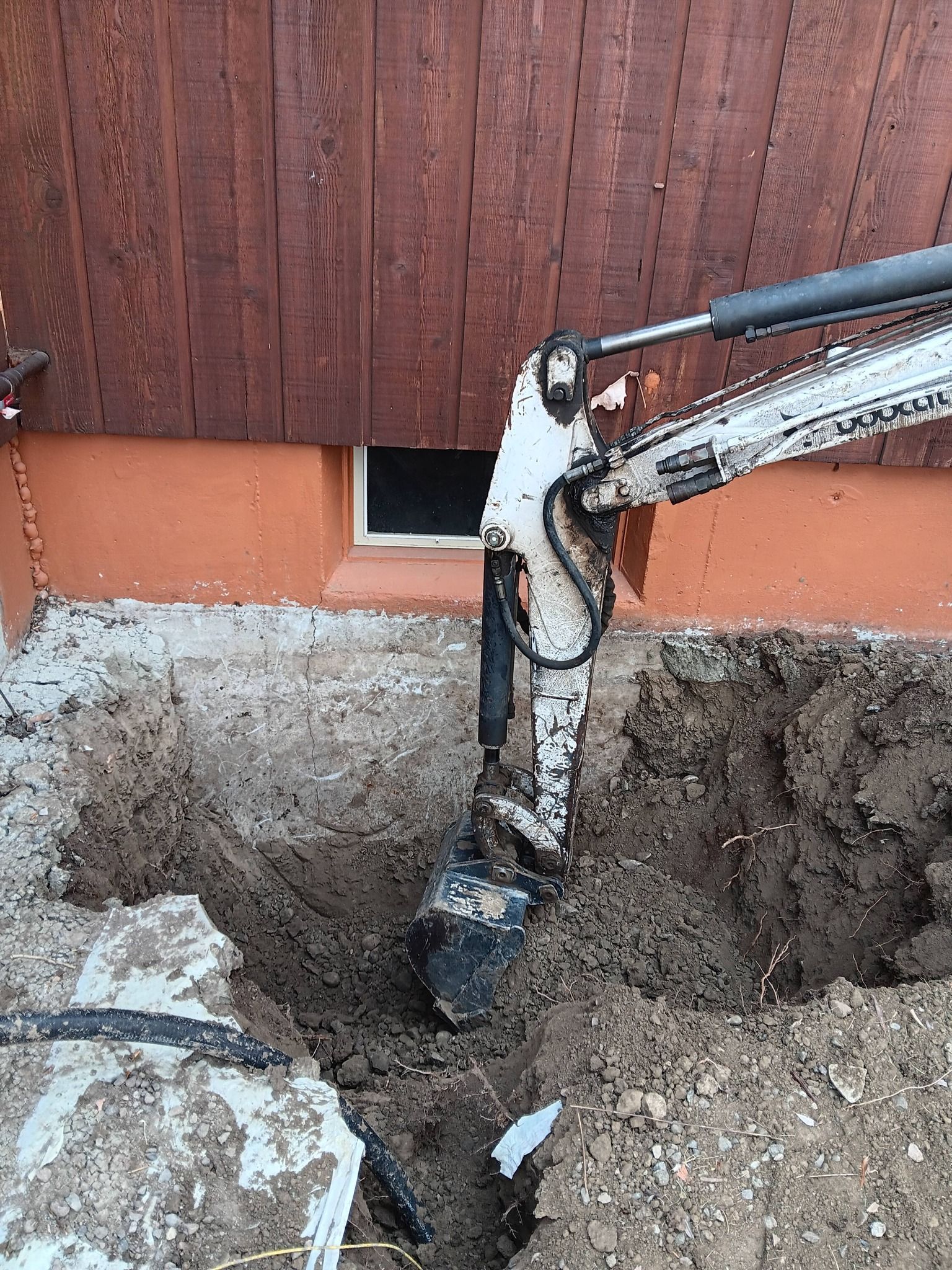 Excavator arm digging a trench beside a house wall with exposed soil and foundation area