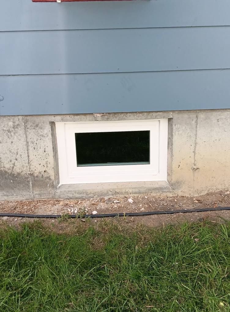 Small basement window in a concrete foundation beneath blue siding, with grass in the foreground.