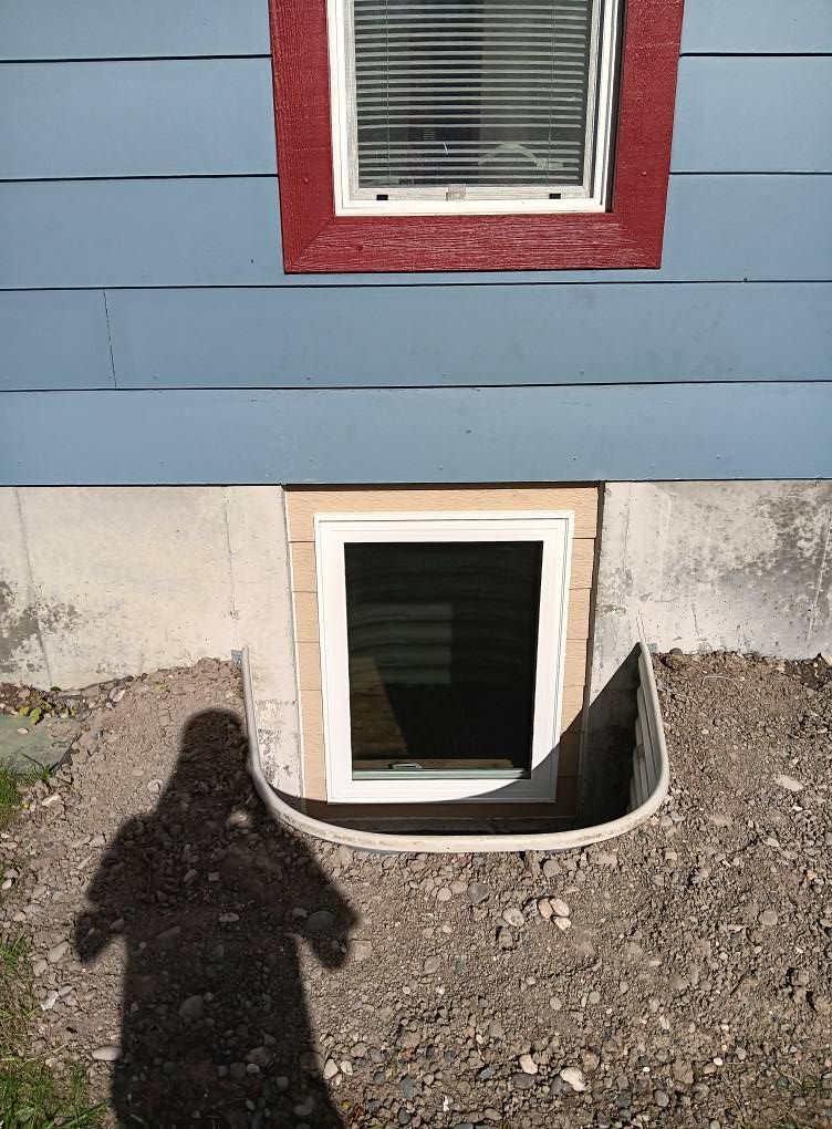 Basement window in a foundation wall below blue siding, with a person’s shadow in the foreground.