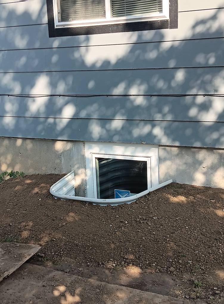 Basement window well beside gray house siding, with dirt and dappled tree shadows.