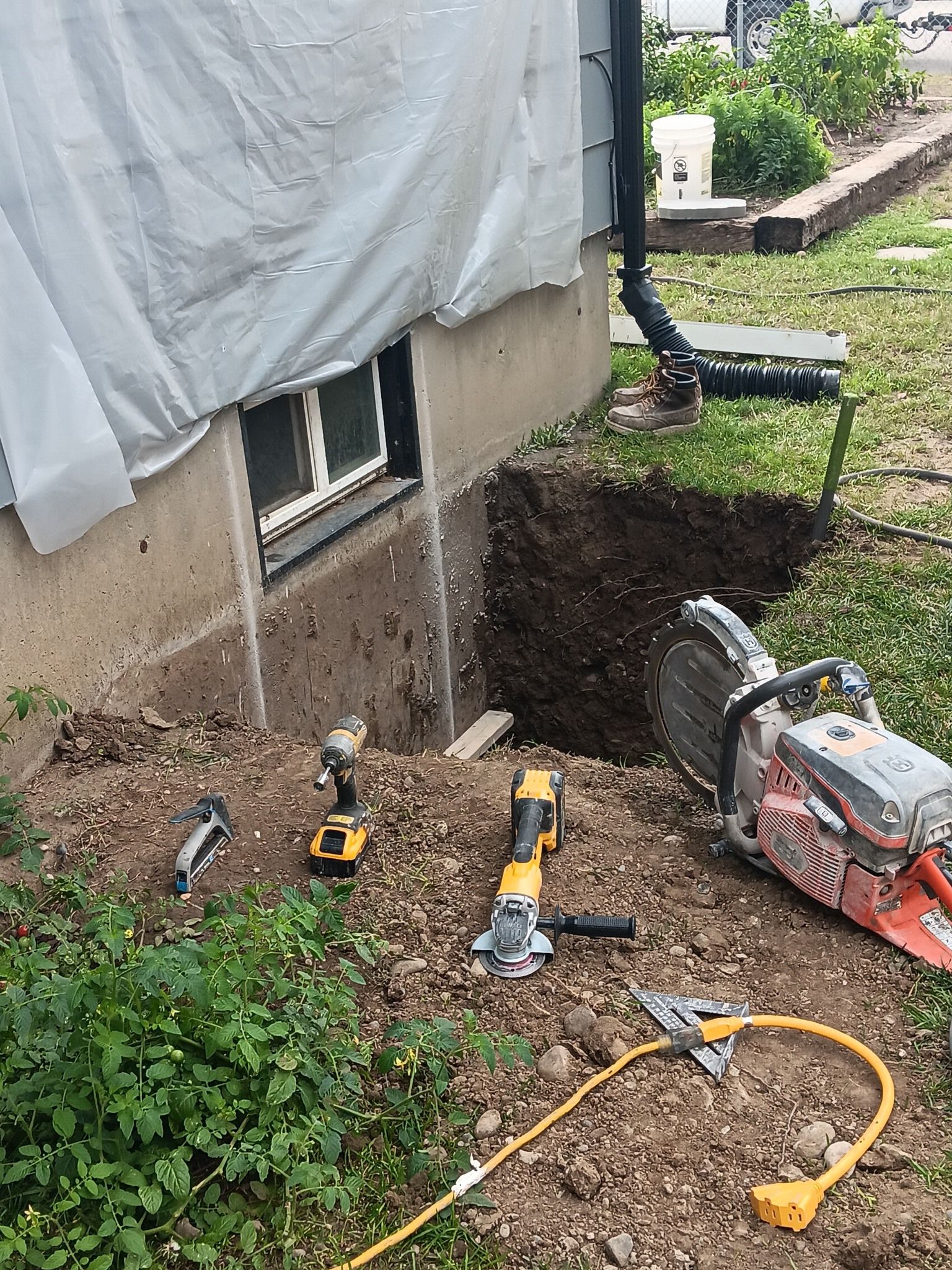 Tools beside a house foundation trench, with a wheelbarrow, shovels, and an extension cord.