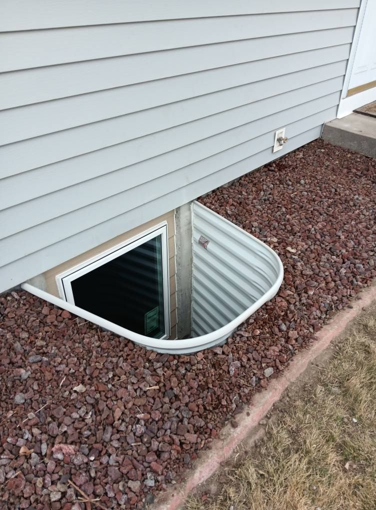 Basement window well with white metal cover beside gray siding and gravel landscaping