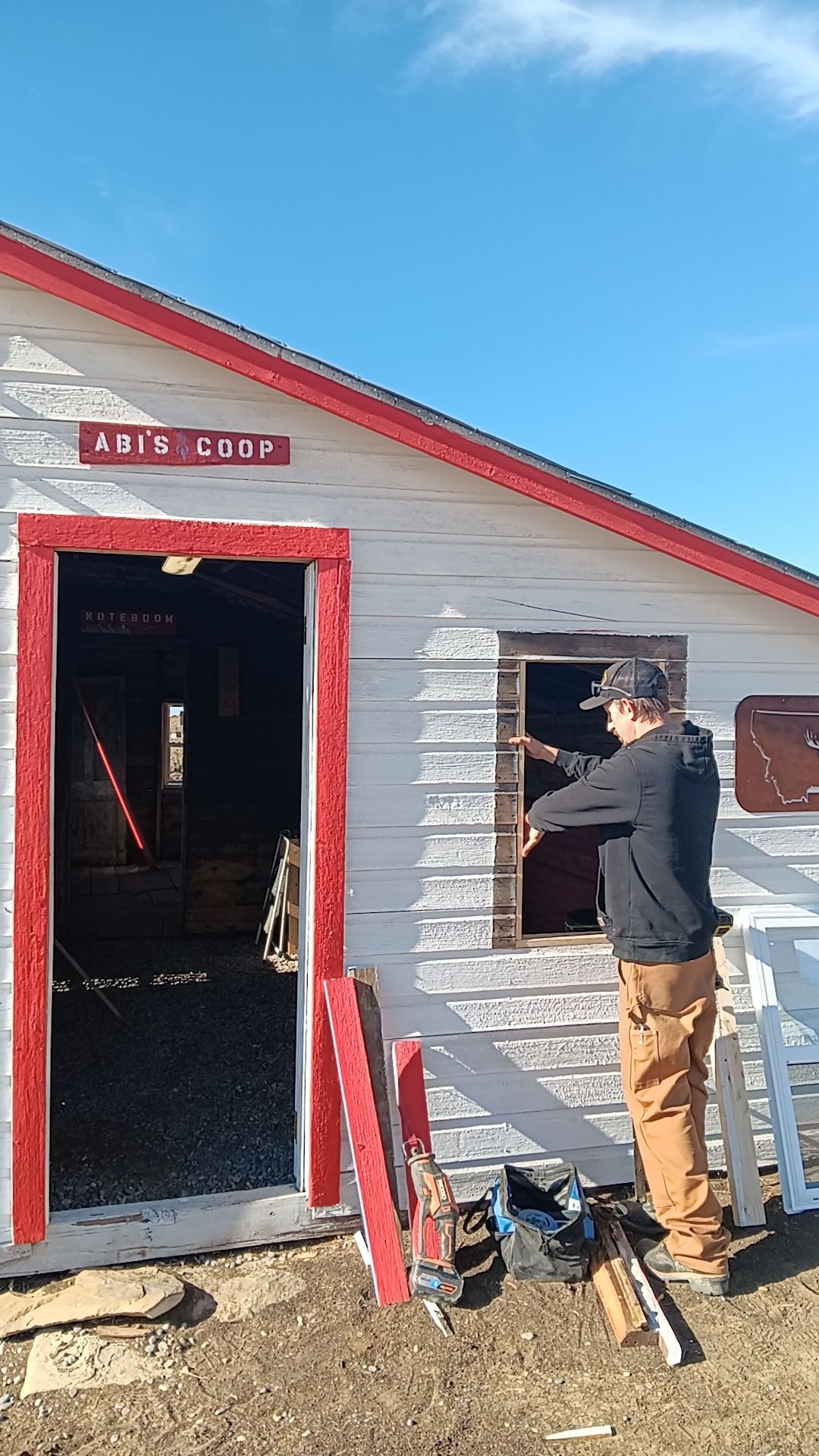 Person in a black jacket works on the side of a small white shed with a red door on a sunny day