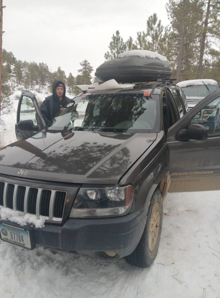 SUV stuck in snowy lot with open doors and roof cargo box, person standing beside it