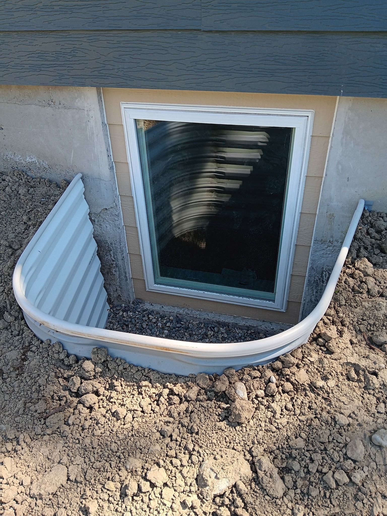 Open basement window with damaged white vent cover on gravel beside house foundation