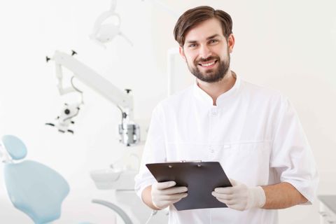 A dentist is smiling while holding a clipboard in a dental office.