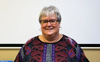 A woman wearing glasses and a purple shirt is smiling in front of a projector screen.
