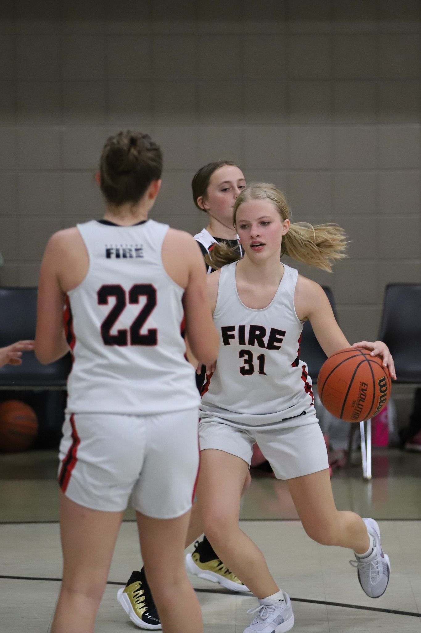 Two women in white basketball uniforms dribble during a game, with another player in the background.