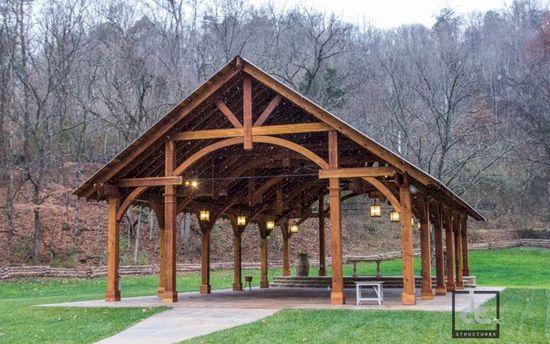 A large, open-air timber-frame pavilion with gabled roof and hanging lights, set in a grassy field backed by trees.