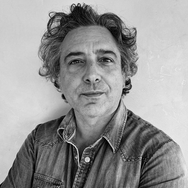 Ritratto di una persona con i capelli ricci, che indossa una camicia di jeans e guarda in avanti, su uno sfondo neutro.