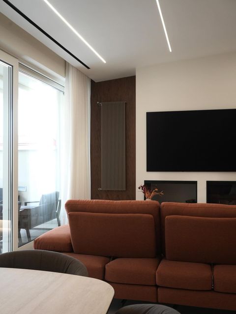 Soggiorno moderno con divano marrone, TV a parete, ampia finestra e lampade a soffitto minimaliste.