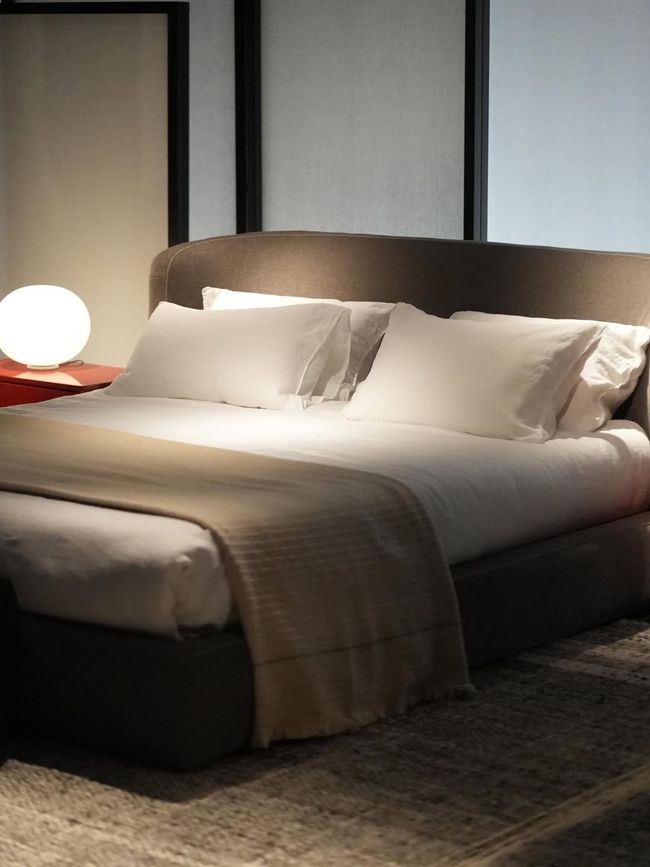 Camera da letto moderna con letto ben rifatto, cuscini bianchi, plaid beige e una lampada da comodino luminosa.